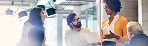 Business people looking at female colleague explaining documents while standing at conference table